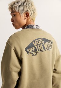 Sudadera de cuello redondo color caqui con un gran gráfico negro que dice "VANS 'OFF THE WALL'" en la parte posterior. La tela parece suave y tiene un corte relajado.