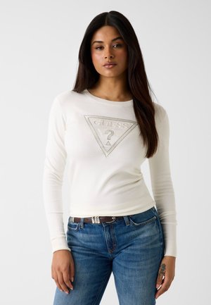 Femme aux longs cheveux foncés portant un pull blanc Guess avec logo et un jean bleu, debout devant un fond clair uni.