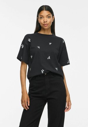 Kvinde iført en sort t-shirt med små hvide blomsterprint og sorte højtaljede bukser, stående mod en ensfarvet hvid baggrund.