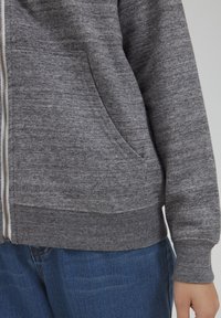 Sivi zip-up dukserica od teksturirane tkanine, s kenguručim džepom, rebrastim manžetama i donjim rubom. Nosena preko plavih traperica.