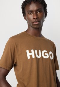 Jaunas vyras su trumpais garbanotais plaukais, vilkintis rudą marškinėlį su balta "HUGO" logotipu, žiūri į kamerą prieš vientisą šviesią fono.
