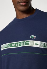 Tamsiai mėlynas medvilninis marškinėliai su žaliu Lacoste logotipu ir raštuotu juosta per krūtinę su baltomis detalėmis ir žaliais siuvinėjimo akcentais.