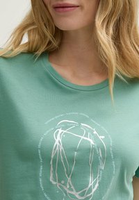 T-shirt vert à manches courtes en coton, avec un col rond et un imprimé abstrait blanc comportant le texte "histoires d'été" en motif circulaire.