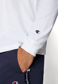 Hvit langermet skjorte med mansjetter, med en liten brodert logo i rødt og svart. Kombinert med marineblå shorts som viser logodetaljer.