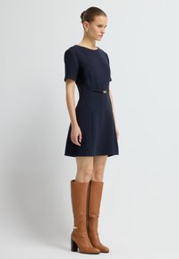 Marine jurk met korte mouwen en een aansluitend ontwerp, met een gouden accent op de taille. Gecombineerd met kniehoge bruine leren laarzen met een chunkyrand.