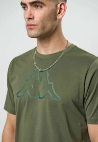 Mann iført olivengrønn t-skjorte med to sittende speilvendte silhuetter som logo og sølvkjede, vendt mot venstre, nøytral bakgrunn.