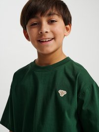 Uśmiechnięty chłopiec o brązowych włosach, ubrany w ciemnozieloną koszulkę z małym beżowym emblematem pszczoły na piersi, na jednolitym jasnym tle.