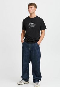 Camiseta de algodón negra con texto gráfico blanco e ilustraciones. Combinada con pantalones de mezclilla de corte holgado y zapatillas deportivas multicolores.