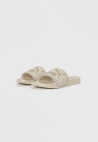 Beige slide sandals with a wide strap featuring the "PUMA" logo and a textured finish. Soft sole for comfort. Minimalist design.