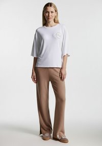 Weißes, kurzärmeliges Hemd mit Rundhalsausschnitt und einem Logo auf der Brust, kombiniert mit hellbraunen, strukturierten, weit geschnittenen Hosen.