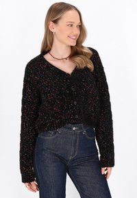 Cardigan nero in tessuto testurizzato con macchie multicolori, scollo a V e chiusura con bottoni; abbinato a jeans blu scuro.