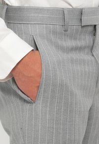 Pantaloni grigi a righe sottili con una texture liscia, dotati di tasche laterali e una vita sagomata. Primo piano di una mano nella tasca.