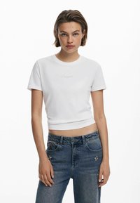 Vit kortärmad t-shirt med figursydd passform och broderad logotyp på bröstet, tillsammans med högmidjade blå jeans med utsmyckade detaljer.