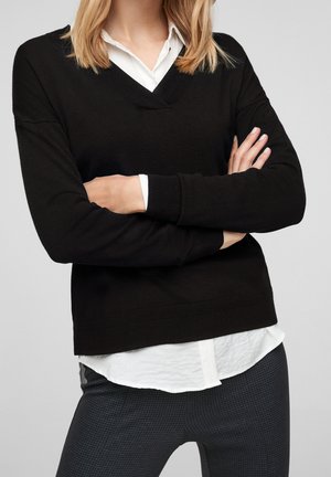 Vrouw draagt een zwart V-hals trui over een wit shirt, met gekruiste armen en donkere, gestructureerde broek tegen een effen lichte achtergrond.