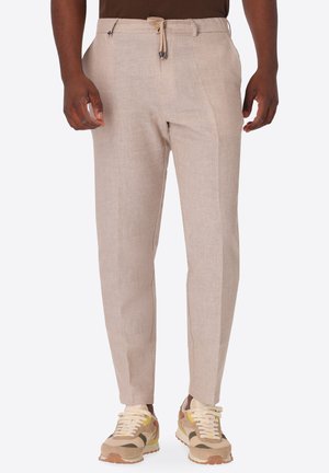 Uomo che indossa pantaloni beige slim-fit con vita con coulisse e sneakers beige multicolore, in piedi su uno sfondo semplice.