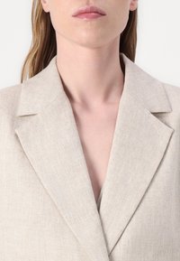 Beige len blézer kiemelkedő gombokkal és texturált anyaggal, mély V-nyakkivágással; letisztult vonalak és minimalisták dizájn. Nincsenek látható gombok vagy díszítések.