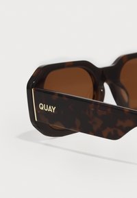 Lunettes de soleil en écaille de tortue avec une monture géométrique, des verres marron et un logo "QUAY" doré sur le côté. Matière en plastique lisse.