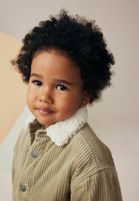 Giacca di corduroy color khaki con un morbido colletto in shearling bianco, caratterizzata da una texture a coste e due bottoni a pressione grigi sul davanti.