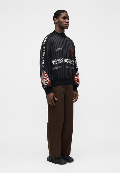 Black knitted sweater with vertical stripes, featuring contrasting red and white text and logo designs. Worn with brown wide-leg trousers.