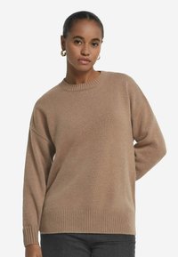 Beiger Strickpullover mit rundem Ausschnitt, gerippten Ärmelbündchen und Saum sowie lässiger Passform. Die Textur wirkt glatt und weich.