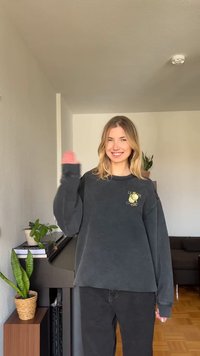 Jeune femme aux cheveux blonds portant un sweat-shirt noir qui fait signe en souriant dans une pièce lumineuse avec des plantes et un piano en arrière-plan.
