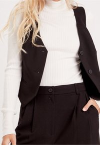 Zwart, op maat gemaakt vest met twee knopen, gedragen over een witte geribbelde coltrui. Zichtbare zakdetails en gestructureerd ontwerp.