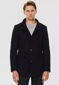 Czarny wełniany płaszcz z kołnierzem, zapinany na guziki z przodu, z dwoma bocznymi kieszeniami i prostym krojem. W parze z jasnoszarym swetrem pod spodem.