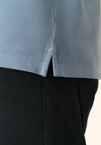 Gros plan d'un ourlet de chemise bleu clair avec une fente latérale sur un pantalon noir, montrant la texture du tissu et les détails des coutures.