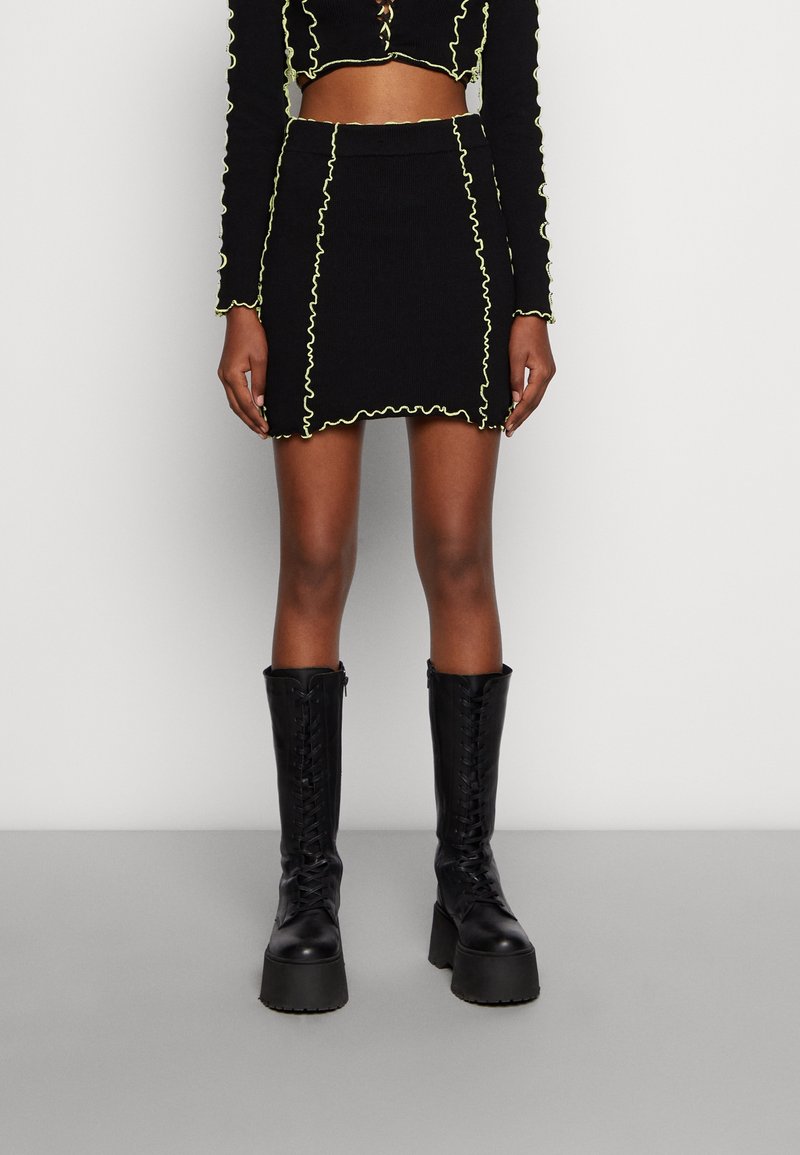 Mini-jupe noire côtelée avec bordure ondulée jaune néon, associée à un crop top assorti et des bottines noires à plateforme lacées.