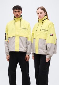 Δύο μοντέλα με κίτρινες και γκρι μπουφάν Tommy Jeans με ψηλούς γιακάδες, μαύρο παντελόνι, στέκονται μπροστά σε μονόχρωμο φόντο.