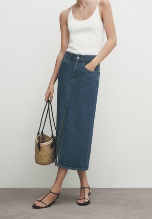 Kobieta w białym topie na ramiączkach, niebieskiej dżinsowej spódnicy o długości do połowy łydki z rozcięciem z przodu, czarnych sandałach na paski, trzymająca plecioną torbę z czarnymi uchwytami.