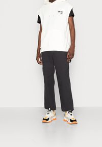 Vit ärmhållen hoodie med ficka fram, svart t-shirt, mörka byxor och flerfärgade sneakers med tjock sula och accenter.