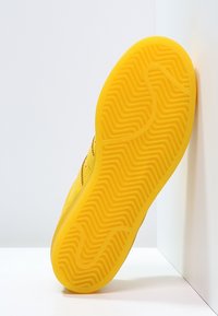 Suela de zapato de goma amarilla con un patrón en espiga texturizado. Presenta un borde liso y una construcción sólida. Exhibida en posición vertical sobre un fondo blanco.