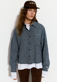 Camicia a quadri bianca e nera in flanella con patta di bottoni, indossata sopra una maglietta a maniche lunghe bianca, abbinata a pantaloni marroni e un cappello marrone.