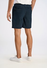 Pantaloni corti in velluto a coste blu navy con vita elastica, tasche laterali e orlo rifinito, indossati con sneakers bianche, con un piccolo tatuaggio visibile sulla gamba.