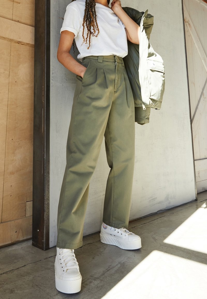 Olijfgroen high-waisted broeken met plooien, gecombineerd met een wit t-shirt en witte platform sneakers. Een groene jas hangt over een arm.
