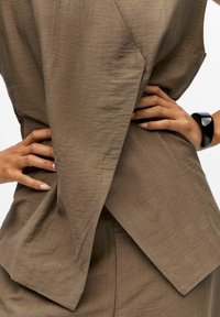 Top marrone e testurizzato con elementi di design angolari e schiena drappeggiata. Bracciale nero evidenzia le mani sui fianchi.