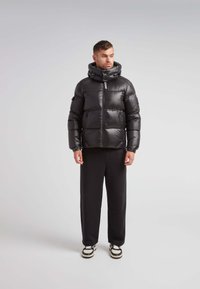 Giacca a vento nera con collo alto, finitura lucida, chiusura con zip e design a pannelli. Abbinata a pantaloni della tuta neri e sneakers.