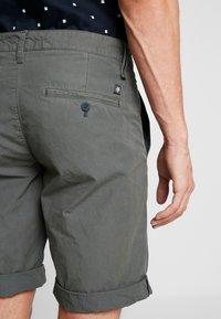 Ingrandimento di una persona che indossa pantaloni corti grigi arrotolati con una tasca posteriore con bottone e una camicia nera a fantasia infilata dentro.