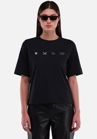 Zwarte katoenen T-shirt met korte mouwen, een losse pasvorm en vier witte bedrukte ontwerpen: zon, vlinder, tekst en een cirkelvormig embleem.