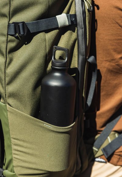 Botella de agua negra en un bolsillo de una mochila de tela verde. La mochila cuenta con correas ajustables y detalles de cremallera. El material parece duradero y texturizado.