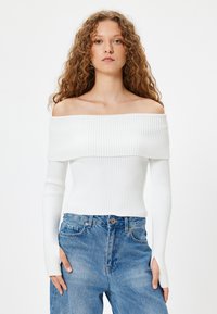 Weißes, schulterfreies, geripptes Oberteil mit langen Ärmeln, figurbetontem Schnitt und strukturiertem Stoff. Kombiniert mit hochtaillierten blauen Jeans.