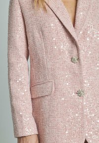 Blazer rosa testurizzato con paillettes, dotato di rever a contrasto, tasca singola e bottoni decorativi a forma di fiore.