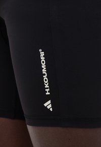 Detailný záber na čierne športové šortky s bielym vertikálnym nápisom "H.KOUMORI" a logom Adidas s tromi pruhmi na stehne.
