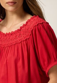 Rote Bluse mit weitem, besticktem Blumen-Ausschnitt, puffärmel und lockerer Passform aus weichem Material.