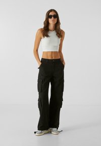 Pantalons cargo noirs avec plusieurs poches, taille haute, coupe large et poignets à cordon, associés avec un crop top blanc et des baskets grises.