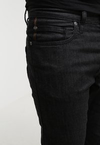 Vaqueros de mezclilla negra con un corte ajustado, diseño de cinco bolsillos, costuras en contraste en color naranja y herrajes metálicos en la cintura y los bolsillos.