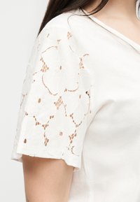 Λευκό μπλουζάκι με κοντά μανίκια και floral δαντελένιο σχέδιο στα μανίκια, φορεμένο από ένα άτομο με μακριά σκούρα μαλλιά που φαίνονται στον ώμο.