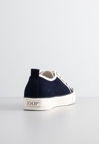 Baskets en toile bleu marine avec semelle en caoutchouc blanche, bout rond et lacets blancs. Présente une étiquette sur le talon avec la marque "JOOP!".