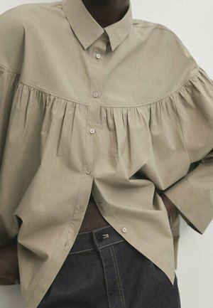 Oversized beige button-up shirt with gathered yoke detail pairs with high-waisted dark denim jeans on a person with one hand on hip.
